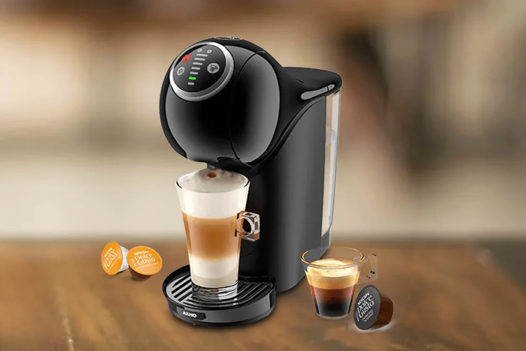 A imagem é composta, da esquerda para a direita, de 2 cápsulas da dolce gusto sob a mesa, ao centro, a cafeteira de cápsula Dolce Gusto Genio S Plus com um copo com bebida a base de leite, e a direita, uma xícara com café e uma cápsula de café sob a mesa de madeira. o fundo está desfocado.