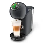 Trata-se de uma imagem que contém a cafeteira de cápsulas Dolce Gusto Genio S Plus de cor preta e fundo branco. Nessa cafeteira, há um copo contendo bebida a base de leite de cor branca e marrom.