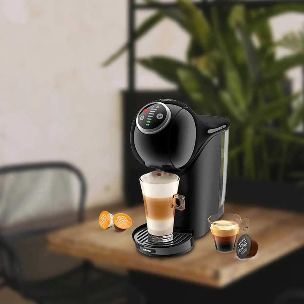 A imagem é composta, da esquerda para a direita, de 2 cápsulas da dolce gusto sob a mesa, ao centro, a cafeteira de cápsula Dolce Gusto Genio S Plus com um copo com bebida a base de leite, e a direita, uma xícara com café e uma cápsula de café sob a mesa de madeira. o fundo está desfocado.