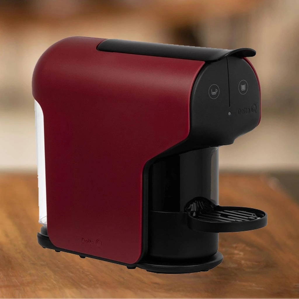 A imagem contém a cafeteira de cápsula Delta Q Quick vista de lateral na cor preta com vermelho. Essa cafeteira está ao centro da imagem e em cima de uma mesa de madeira com o fundo desfocado.