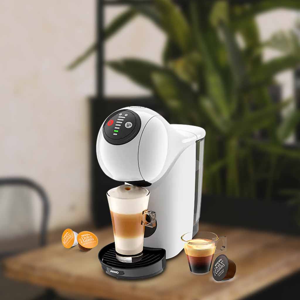 A imagem é composta, da esquerda para a direita, de 2 cápsulas da dolce gusto sob a mesa, ao centro, a cafeteira de cápsula Dolce Gusto Genio S Basic de cor branca com um copo com bebida a base de leite, e a direita, uma xícara com café e uma cápsula de café sob a mesa de madeira. o fundo está desfocado.