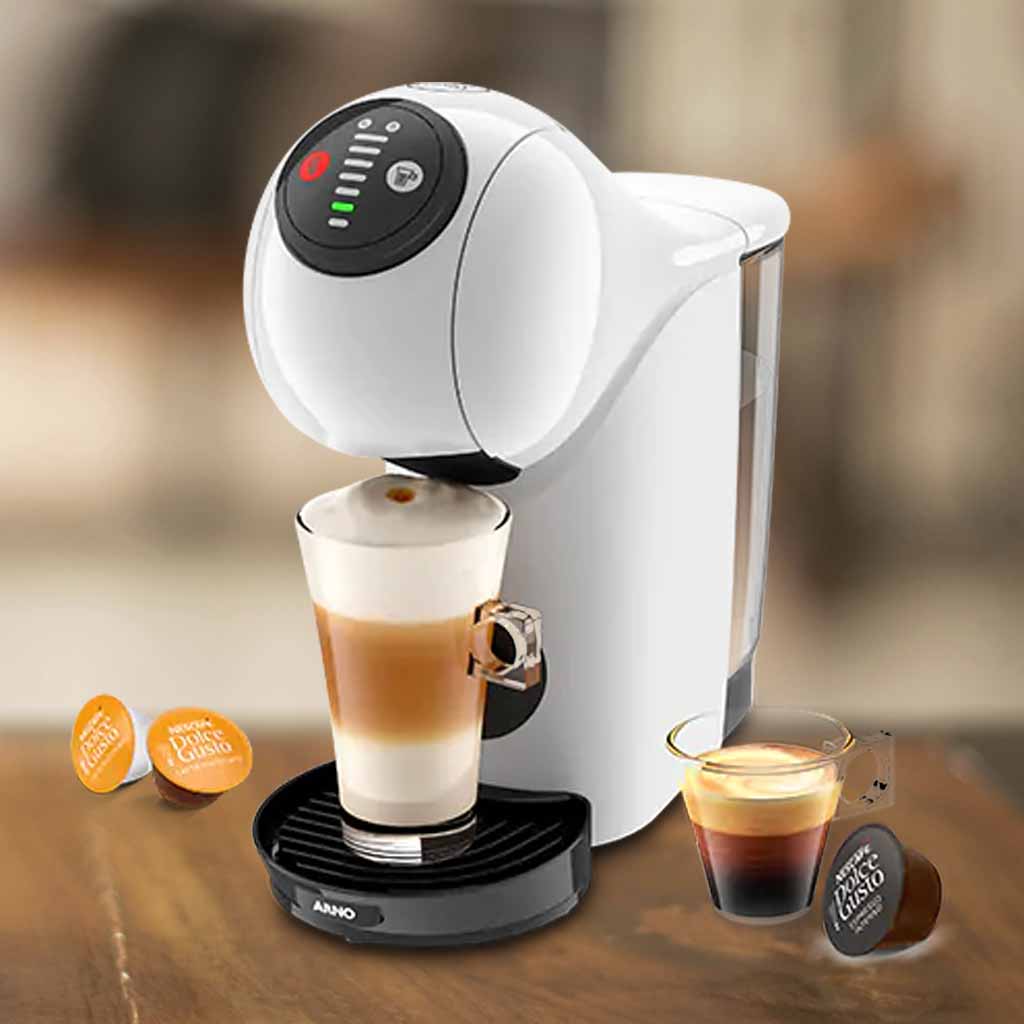 A imagem é composta, da esquerda para a direita, de 2 cápsulas da dolce gusto sob a mesa, ao centro, a cafeteira de cápsula Dolce Gusto Genio S Basic de cor branca com um copo com bebida a base de leite, e a direita, uma xícara com café e uma cápsula de café sob a mesa de madeira. o fundo está desfocado.