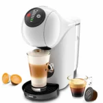 A imagem é composta, da esquerda para a direita, de 2 cápsulas da dolce gusto sob a mesa, ao centro, a cafeteira de cápsula Dolce Gusto Genio S Basic de cor branca com um copo com bebida a base de leite, e a direita, uma xícara com café e uma cápsula de café. Fundo branco.