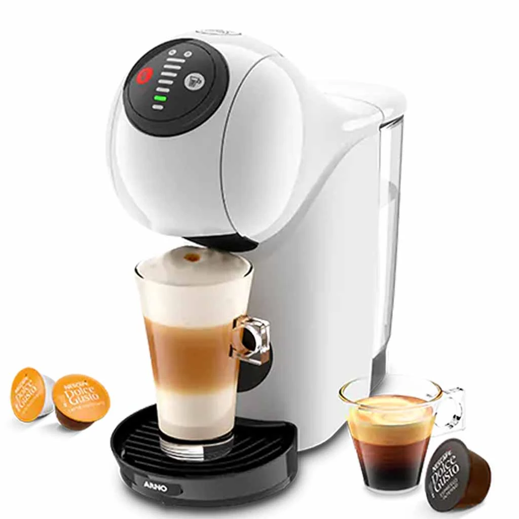 A imagem é composta, da esquerda para a direita, de 2 cápsulas da dolce gusto sob a mesa, ao centro, a cafeteira de cápsula Dolce Gusto Genio S Basic de cor branca com um copo com bebida a base de leite, e a direita, uma xícara com café e uma cápsula de café. O fundo da imagem é branco.