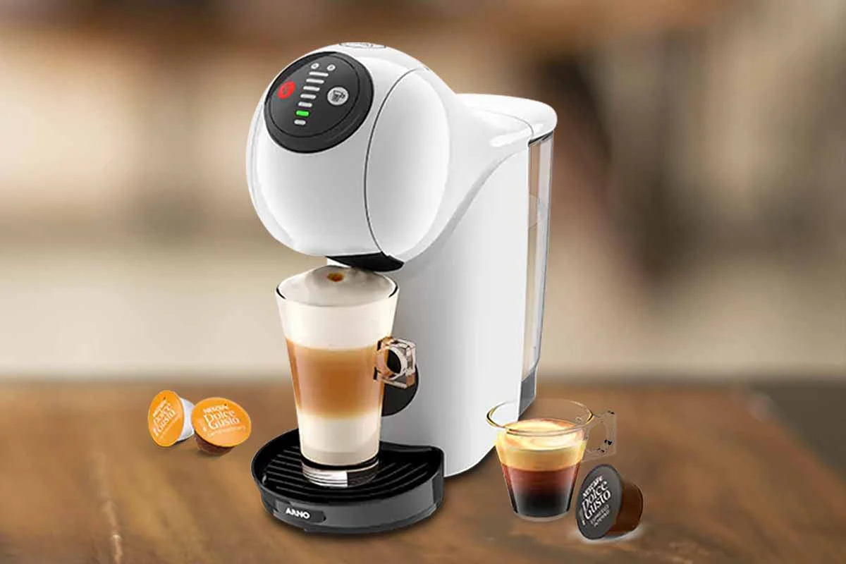 A imagem é composta, da esquerda para a direita, de 2 cápsulas da dolce gusto sob a mesa, ao centro, a cafeteira de cápsula Dolce Gusto Genio S Basic de cor branca com um copo com bebida a base de leite, e a direita, uma xícara com café e uma cápsula de café sob a mesa de madeira. o fundo está desfocado.