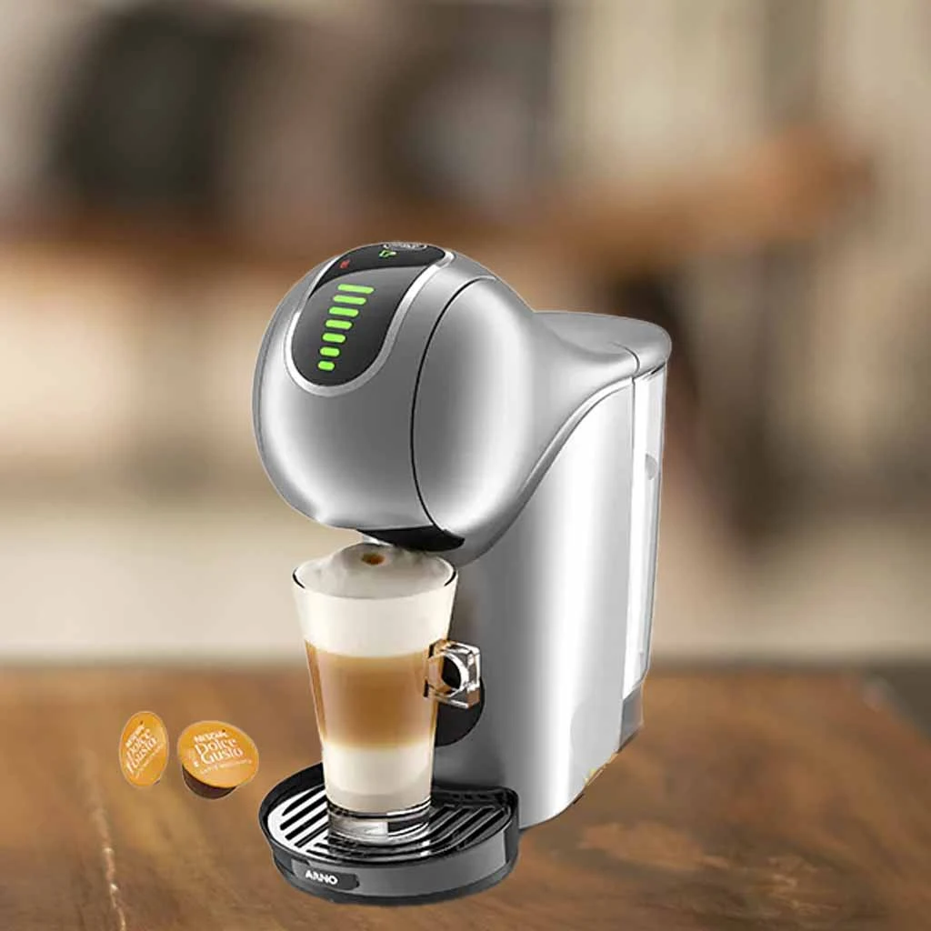 A imagem é composta pela cafeteira de cápsula Dolce Gusto Genio S Touch de cor prata ao centro. Além disso, nessa cafeteira tem um copo com bebida a base de leite dentro. Essa cafeteira está em cima de uma mesa de madeira e o fundo restante está desfocado. À esquerda têm duas capsulas da dolce gusto sob a mesa