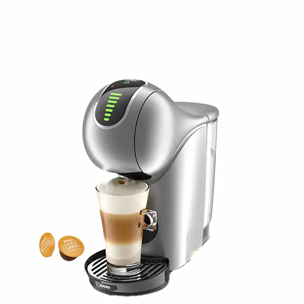 A imagem é composta pela cafeteira de cápsula Dolce Gusto Genio S Touch de cor prata ao centro. O fundo da imagem é branco. À esquerda têm duas capsulas da dolce gusto sob a mesa