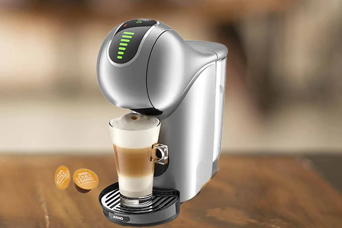 A imagem é composta pela cafeteira de cápsula Dolce Gusto Genio S Touch de cor prata ao centro. Além disso, nessa cafeteira tem um copo com bebida a base de leite dentro. Essa cafeteira está em cima de uma mesa de madeira e o fundo restante está desfocado. À esquerda têm duas capsulas da dolce gusto sob a mesa