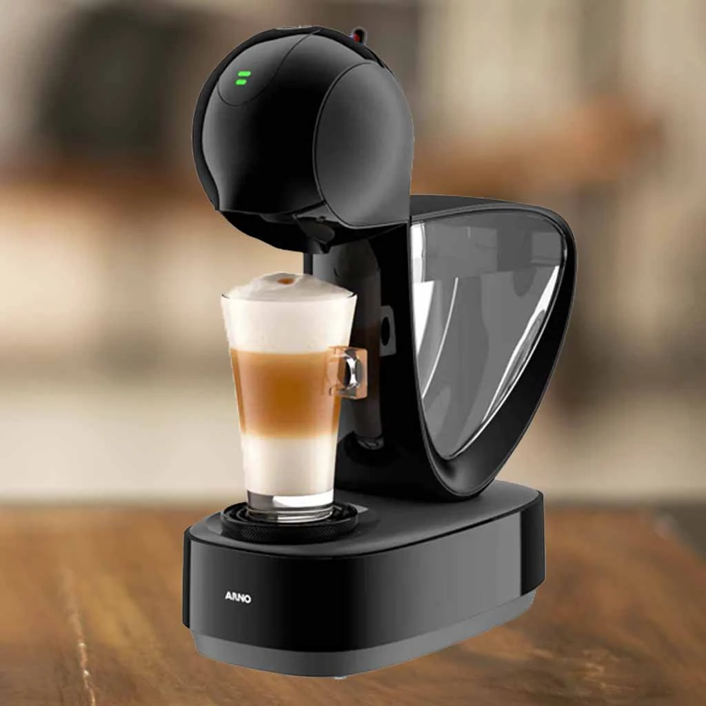 A imagem é composta pela cafeteira de cápsula Dolce Gusto Infinissima de cor preta ao centro. Além disso, nessa cafeteira tem um copo com bebida a base de leite dentro. Essa cafeteira está em cima de uma mesa de madeira e o fundo restante está desfocado.