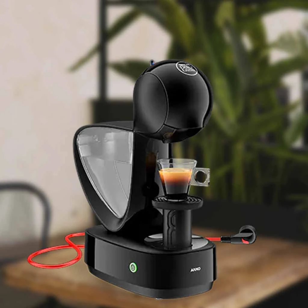 A imagem é composta pela cafeteira de cápsula Dolce Gusto Infinissima de cor preta ao centro. Além disso, nessa cafeteira tem um xícara com café. Essa cafeteira está em cima de uma mesa de madeira e o fundo restante está desfocado.