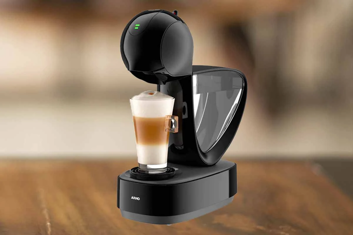 A imagem é composta pela cafeteira de cápsula Dolce Gusto Infinissima de cor preta ao centro. Além disso, nessa cafeteira tem um copo com bebida a base de leite dentro. Essa cafeteira está em cima de uma mesa de madeira e o fundo restante está desfocado.