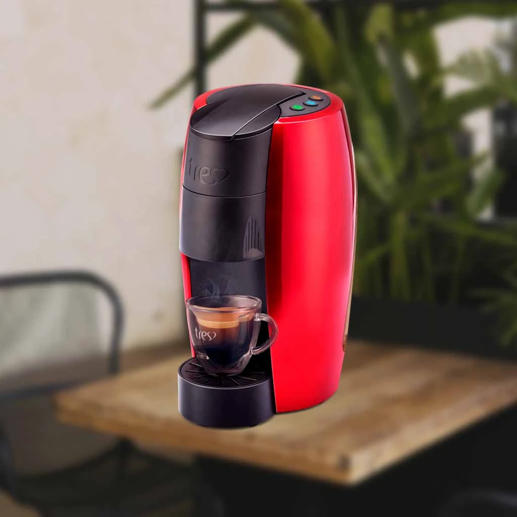 A imagem é composta pela cafeteira de cápsula Três Corações Lov de cor preta com vermelho ao centro da imagem. Essa cafeteira está sob uma mesa de madeira e o restante do fundo é desfocado com plantas e uma cadeira. Há nessa cafeteira uma xícara com café expresso.