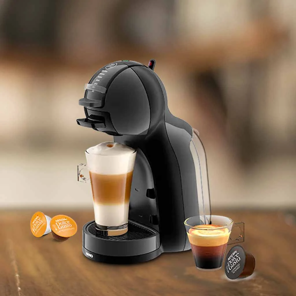 A imagem é composta, da esquerda para a direita, de 2 cápsulas da dolce gusto sob a mesa, ao centro, a cafeteira de cápsula Dolce Gusto MIni Me com um copo com bebida a base de leite, e a direita, uma xícara com café e uma cápsula de café sob a mesa de madeira. o fundo está desfocado.