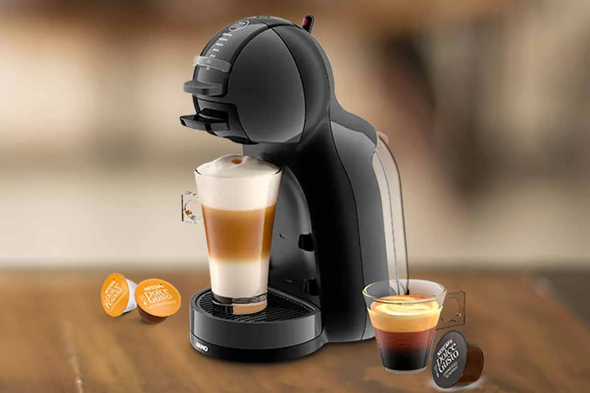 A imagem é composta, da esquerda para a direita, de 2 cápsulas da dolce gusto sob a mesa, ao centro, a cafeteira de cápsula Dolce Gusto MIni Me com um copo com bebida a base de leite, e a direita, uma xícara com café e uma cápsula de café sob a mesa de madeira. o fundo está desfocado.