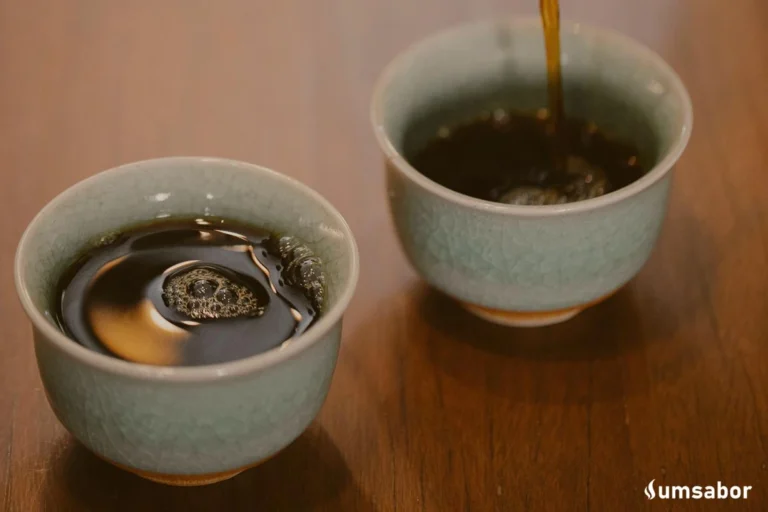 A imagem contém 2 xícaras sob uma mesa com líquido preto (café) nelas. No canto direito inferior há o logo do site um sabor na cor branca