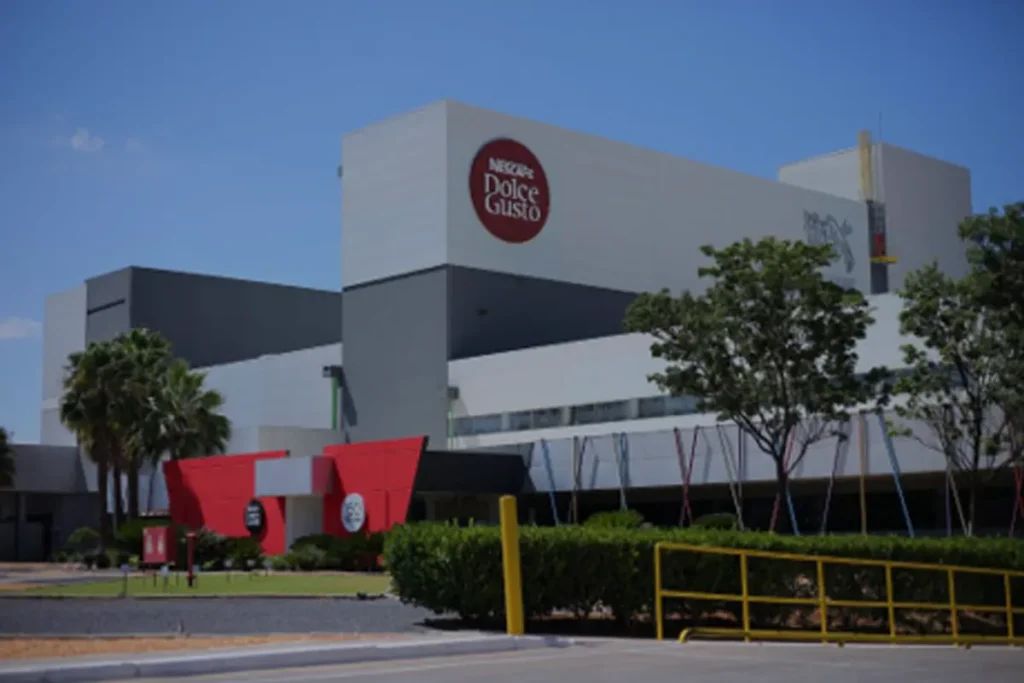 R$ 450 MILHÕES e Mais Café! Nestlé faz o MAIOR investimento da história da Dolce Gusto no Brasil. 2 Vista externa diurna da grande e moderna fábrica da Nestlé Nescafé Dolce Gusto em Montes Claros (MG), uma das cinco unidades da marca no mundo. O edifício apresenta design industrial limpo com predomínio de cores cinza e branco.